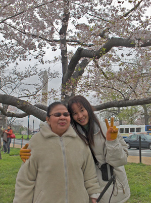 Nanta and Ann stand in front of a cherry tree, with a street visible behind them and in the distance is the Washington Monument.  Ann has one arm around Nanta and with the other hand holds up two fingrs in the 'V for victory' sign.